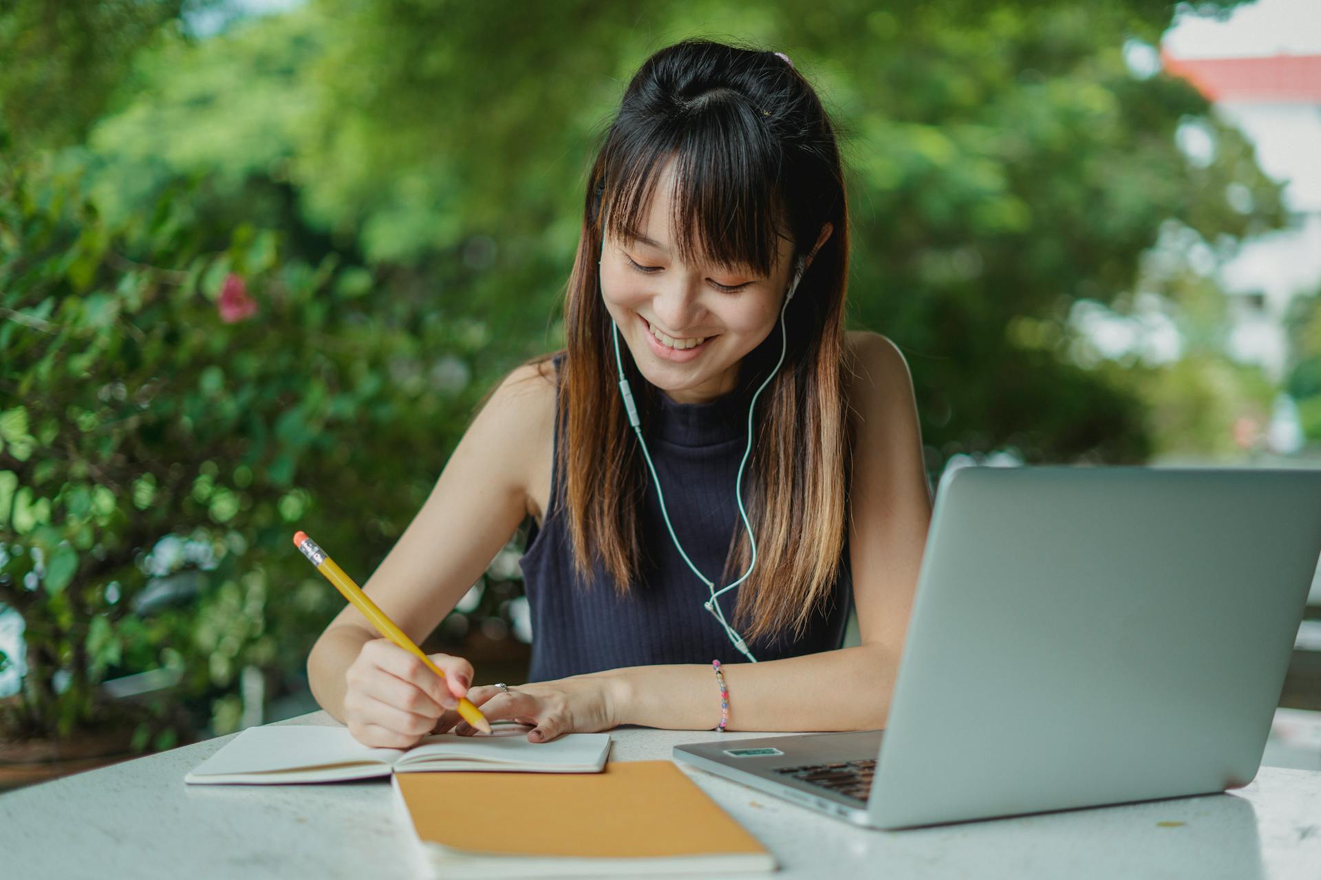 Smiling woman with earphones taking notes beside a laptop outdoors