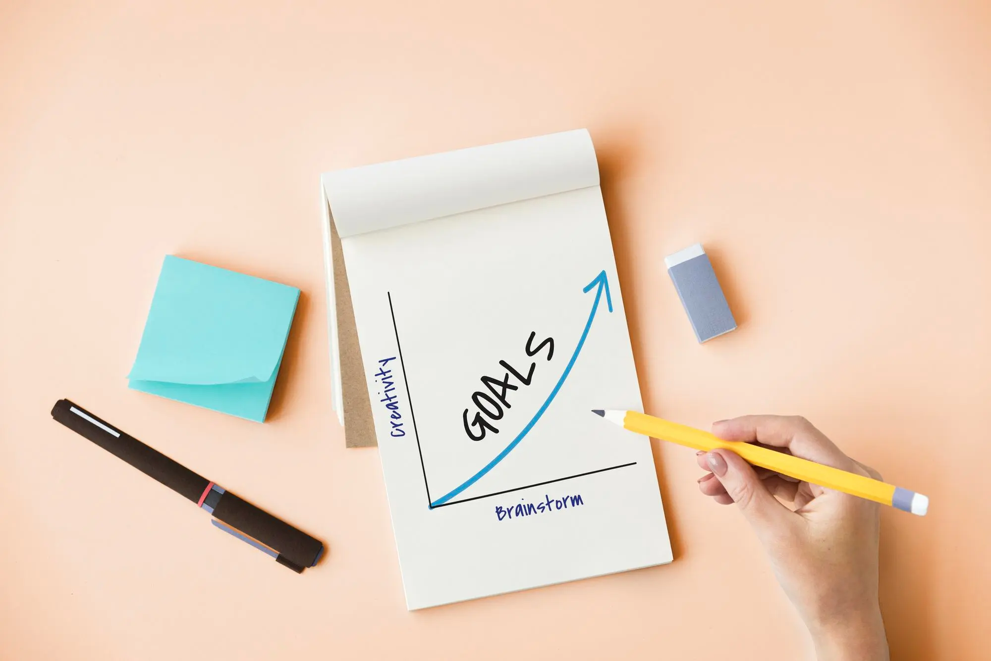 Hand drawing a rising Goals chart on a notepad beside sticky notes and a pen